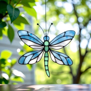 Op de afbeelding: Een gebrandschilderde glazen libelle zonnevanger met blauwe en witte vleugels, een groene body en zwarte accenten. De libelle hangt aan een dun zwart draadje, met een vage groene en witte achtergrond.