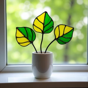 May include: Three stained glass leaves in a white ceramic pot. The leaves are green and yellow with black outlines.