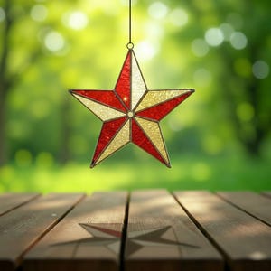 May include: A stained glass star with red, white, and gold segments hangs against a blurred green background. The star is suspended by a thin black wire, casting a shadow on a wooden surface below. The star's design is reminiscent of a patriotic theme.