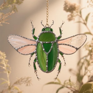 Puede incluir: Un atrapasoles de escarabajo de vidrieras con una cadena dorada para colgar. El escarabajo presenta un cuerpo verde, alas rosas y transparentes, y un marco de metal plateado. El insecto tiene patas y antenas detalladas.