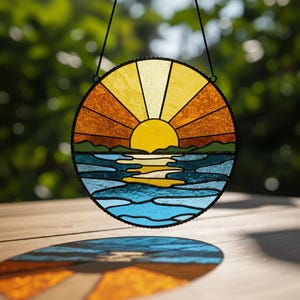 May include: Round stained glass panel depicting a sunset over water. The sun is a bright yellow, with orange and yellow rays. The water is blue and the sky is a gradient of yellow and orange. The panel is framed in black and hangs from a black cord.