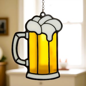 May include: A stained glass beer mug hanging ornament with a yellow beer and white foam. The mug has a black outline and a handle.