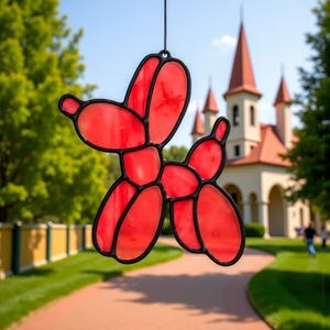 May include: A red stained glass balloon dog with black outlines hangs in front of a blurred background of a building and trees. The balloon dog is a whimsical decoration, perfect for a child's room or as a unique gift.