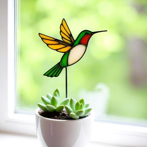 Puede incluir: Un colibrí de vidrio coloreado con detalles amarillos, verdes y rojos está posado en una estaca de metal negro. El colibrí está delante de una planta suculenta en maceta.