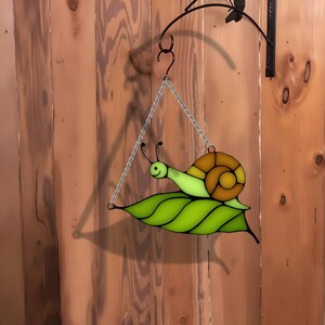 Puede incluir: Un caracol de vidrio coloreado sobre una hoja verde, colgado de una cadena. El caracol es marr&oacute;n y amarillo con un cuerpo verde. La hoja es verde con un centro verde claro.