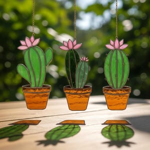 May include: Three stained glass cactus ornaments with pink flowers and orange pots hang from gold chains. The green cacti have varying shapes and are set against a blurred green background. The ornaments cast shadows on a wooden surface.