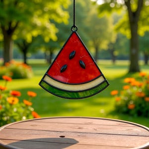 Può includere: Un acchiappasole a forma di fetta di anguria in vetro colorato, con bordo nero, centro rosso, semi neri, scorza bianca e gialla e scorza esterna verde. L'acchiappasole è appeso davanti a uno sfondo verde sfocato.