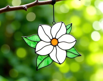 May include: A stained glass flower with white petals, green leaves, and an orange center hangs from a tree branch. The flower is outlined in black and suspended by a black wire. The background is a blurred green.