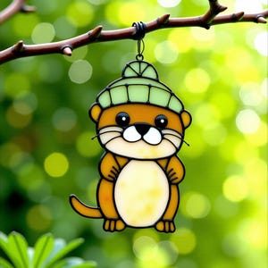 Pode incluir: Um ornamento de vidro manchado de lontra com um chapéu verde claro, pendurado em um galho de árvore. A lontra tem um corpo marrom, rosto branco e olhos pretos. O fundo é verde desfocado com bokeh.