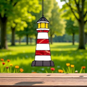 May include: A stained glass lighthouse ornament, featuring alternating red and white stripes, a black roof, and a yellow light. The lighthouse is suspended against a blurred background of green trees and grass.