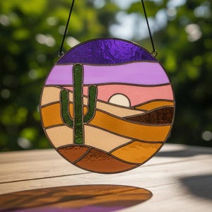 Op de afbeelding: Rond gebrandschilderd glaskunstwerk met een woestijnlandschap. Het toont een groene cactus, een ondergaande zon en gelaagde heuvels in tinten oranje, bruin en roze. De lucht is paars en lavendel. Het stuk is omlijst in donker metaal.