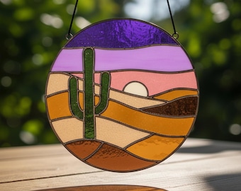 May include: Round stained glass art piece featuring a desert landscape with a green cactus, a setting sun, and layered hills in shades of orange, brown, and pink. The sky is purple and lavender. The piece is framed in a dark metal.