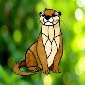 Pode incluir: Estatueta de lontra em vitral com detalhes castanhos, bege e pretos. A lontra está sentada em pé com a cauda enrolada. A estatueta tem um fio preto para pendurar e está sobre um fundo verde desfocado.