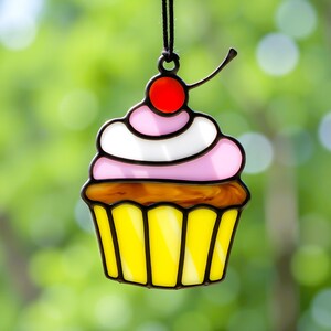 May include: A stained glass cupcake ornament with pink and white frosting, a red cherry on top, and a yellow cupcake liner.
