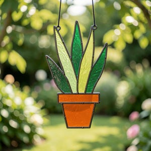 Op de afbeelding: Een glas-in-lood suncatcher in de vorm van een potplant met puntige bladeren in verschillende tinten licht- en donkergroen. De plant staat in een oranje pot met een donker metalen frame dat de glasstukken bij elkaar houdt, opgehangen aan een zwart koord.