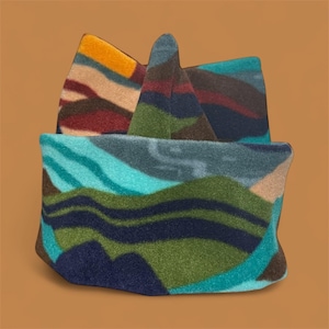 May include: A patterned beanie hat with a landscape design. The hat features a mountain range design in shades of blue, green, brown, and yellow. The hat is made of a soft, textured material.