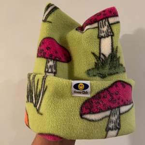 May include: A light green fleece beanie with a repeating pattern of red and white mushrooms. The beanie has a folded brim and a small tag that reads "Snow Club" with a smiling face logo. The hat is designed for cold weather.