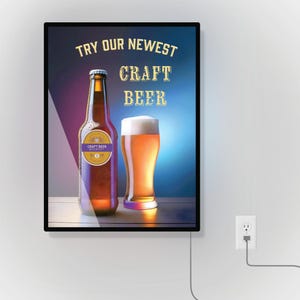 May include: A black framed sign with the text "TRY OUR NEWEST CRAFT BEER" in gold lettering. A bottle of beer with a label that says "CRAFT BEER BREWERY" and a glass of beer are in front of a blue and purple background.