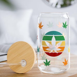 May include: A clear glass can-shaped drinking glass with a bamboo lid and straw. The glass features a colorful design of cannabis leaves and a circular graphic with a cannabis leaf in the center.
