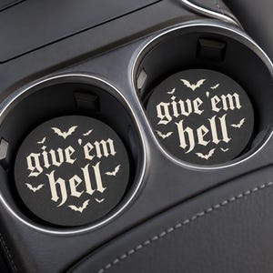 Puede incluir: Dos posavasos circulares negros para portavasos de coche con el texto blanco give 'em hell en fuente gótica, rodeado de pequeñas siluetas de murciélagos blancos. Los posavasos están colocados dentro de los portavasos de una consola.