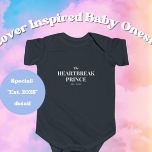 May include: A dark gray baby onesie with the text "The HEARTBREAK PRINCE EST. 2025". The words "Lover Inspired Baby Onesie" are arched above the onesie. A light blue circle reads "Special: 'Est. 2025' detail".