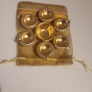 May include: A collection of seven gold-coloured oil lamps, arranged in a circular pattern, presented on a gold-coloured sheer fabric pouch. The lamps feature a traditional design with a central bowl and a spout for the wick. The pouch has decorative gold trim.