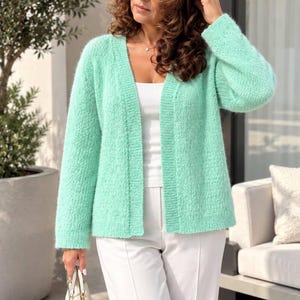May include: A mint green open-front cardigan with a textured knit pattern, long sleeves, and a ribbed trim along the neckline and front edges. The garment is worn over a white top and white trousers, paired with a small white handbag.