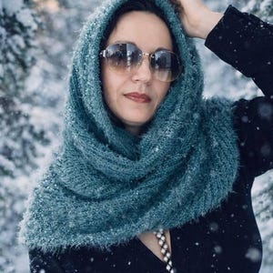 May include: A woman wearing a teal knitted cowl scarf and sunglasses in a snowy outdoor setting. The scarf is wrapped around the head and neck, providing warmth and style. The background features snow-covered trees.