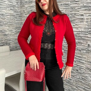May include: A woman wearing a red cardigan with a black lace top and black trousers. She is holding a red ostrich leather handbag.