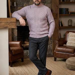May include: A man wearing a lavender cable-knit sweater, dark gray trousers, and brown leather shoes. The sweater has a crew neck and a textured pattern. The trousers are a solid color, and the shoes are laced. The man is standing in a room with a fireplace and leather chairs.