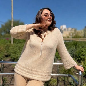 May include: A woman wearing a beige sweater with a decorative neckline and matching beige pants. The sweater has long sleeves and a ribbed hem. She is wearing sunglasses and a necklace with a long tassel. The background is a park with a blue sky.