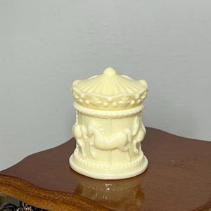 May include: A miniature carousel figurine in a soft yellow color. The figurine features detailed relief carvings of horses and decorative elements. The carousel has a domed top and a circular base, creating a charming, whimsical aesthetic.