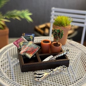 May include: Miniature gardening set featuring a dark brown wooden tray, small terracotta pots, seed packets, and gardening tools. The set includes a small tree, grass, and a trowel. The tray sits on a white metal table.