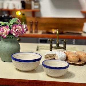 May include: A miniature kitchen scene featuring a green vase with pink and white flowers, two white bowls with blue rims, and a wooden board with bread and cheese. The scene is set on a light-coloured surface.