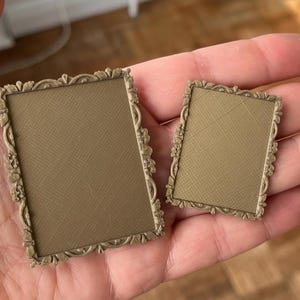 May include: Two small, ornate, rectangular picture frames with a textured, tan surface. The frames have a decorative, floral border in a muted gold colour. One frame is larger than the other.