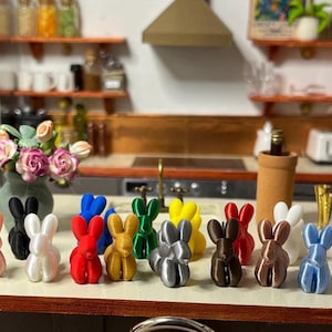 May include: A selection of miniature bunny figurines in a range of colours, including pink, black, white, red, gold, green, yellow, grey, brown, and blue. The bunnies are displayed on a countertop in a kitchen.