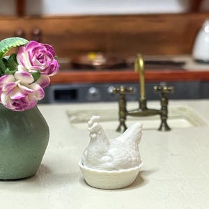 May include: A small white hen figurine in a basket. A green vase with pink and white flowers is to the left. A brass faucet is in the background, suggesting a kitchen setting. The scene is a miniature display.