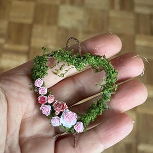 May include: A miniature heart-shaped wreath, featuring a moss base and a collection of tiny pink and red roses. The wreath is held in a hand, highlighting the intricate details of the floral design.