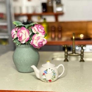 May include: A miniature scene featuring a small, pale green vase with pink and white roses. A white teapot with a floral wreath design sits nearby. The teapot has a gold-coloured knob on the lid. The scene is set on a light-coloured surface.