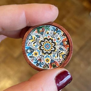 May include: A round wooden coaster with a colourful floral design. The coaster features a central mandala with blue, yellow, red, and green flowers. The coaster is perfect for protecting surfaces from heat and spills.