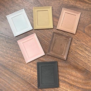 May include: Six small rectangular picture frames in a variety of colours: white, gold, peach, pink, brown, and black. The frames are displayed on a wooden surface, highlighting their different shades and textures.