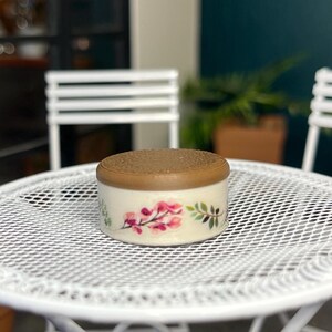 May include: A small, round, decorative box with a light beige base and a brown lid. The box is adorned with a floral design featuring pink flowers and green leaves. The box is sitting on a white, metal, mesh table.