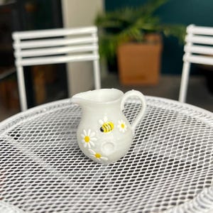 May include: A miniature white ceramic jug with a handle and spout, adorned with yellow and black bees and white daisy flowers. It rests on a white, metal mesh table. White chairs are visible in the background.