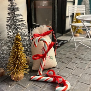 May include: Miniature Christmas scene with a small burlap sack tied with a red ribbon, and two candy canes with red and white stripes and a touch of blue. A small gold and silver Christmas tree is in the background.