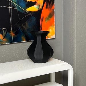 May include: A small, black vase with a rounded body and flared rim. The vase is on a white shelf, with a colourful abstract painting in the background. The vase has a glossy finish and a simple, elegant design.