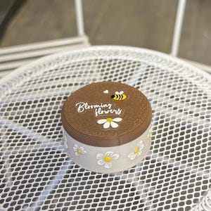 May include: A round container with a brown lid and a grey base. The lid has the text "Blooming Flowers", a bee and a daisy. The base is decorated with white daisies with yellow centres.