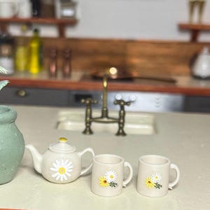 May include: Miniature cream tea set with daisy designs, including a teapot and two mugs. Ideal for a doll's house kitchen. The teapot has a yellow knob. The mugs and teapot feature a white daisy design with yellow centres.