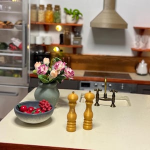 May include: A miniature kitchen scene featuring a gray bowl of red fruit, a vase of pink flowers, and two wooden salt and pepper shakers. A refrigerator and shelves with jars and plants are in the background.