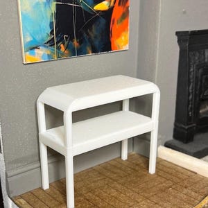 May include: A small, white two-tiered bench with curved edges. The bench has a textured surface, suitable for a doll's house or miniature scene. It's positioned against a grey wall, with a colourful abstract painting above.