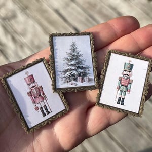 May include: Three miniature framed Christmas-themed prints. One shows a snow-covered Christmas tree with gifts, and the other two depict nutcracker soldiers in pink and green uniforms. The frames have an ornate, antique gold finish.